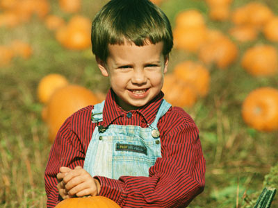 A child in a red and black striped shirt and blue overalls holding up a pumpkin.